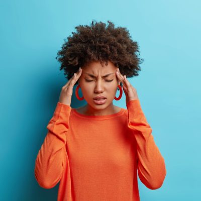 Photo of frustrated young Afro American woman has headache keeps hands on temples suffers unbearable migraine after noisy party wears orange sweater poses indoor isolated on blue background. Photo of frustrated young Afro American woman has headache keeps hands on temples suffers unbearable migraine after noisy party wears orange sweater poses indoor isolated on blue background.